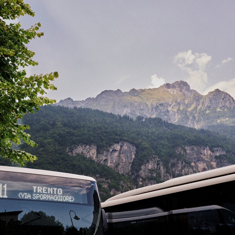 Parte superiore anteriore di un autobus con il display luminoso che indica "B611 TRENTO (VIA SPORMAGGIORE)". Dietro il veicolo si erge un'imponente montagna rocciosa sopra pendii coperti di alberi. | © Origami, 2026