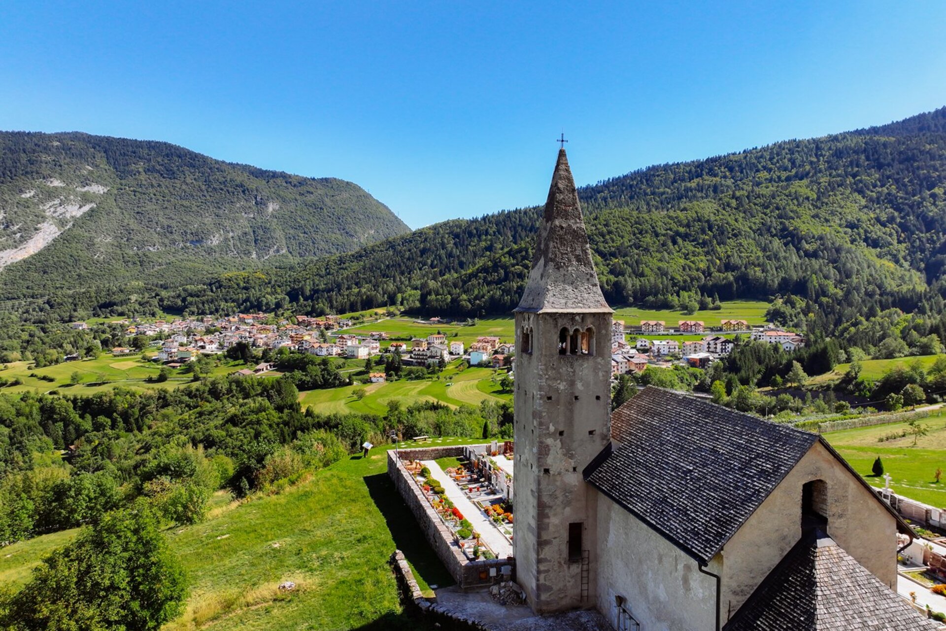In primo piano c'è la chiesetta di San Tommaso di Cavedago, dietro si vedono alcune case del paese e sullo sfondo i boschi di sempreverdi | © 2022