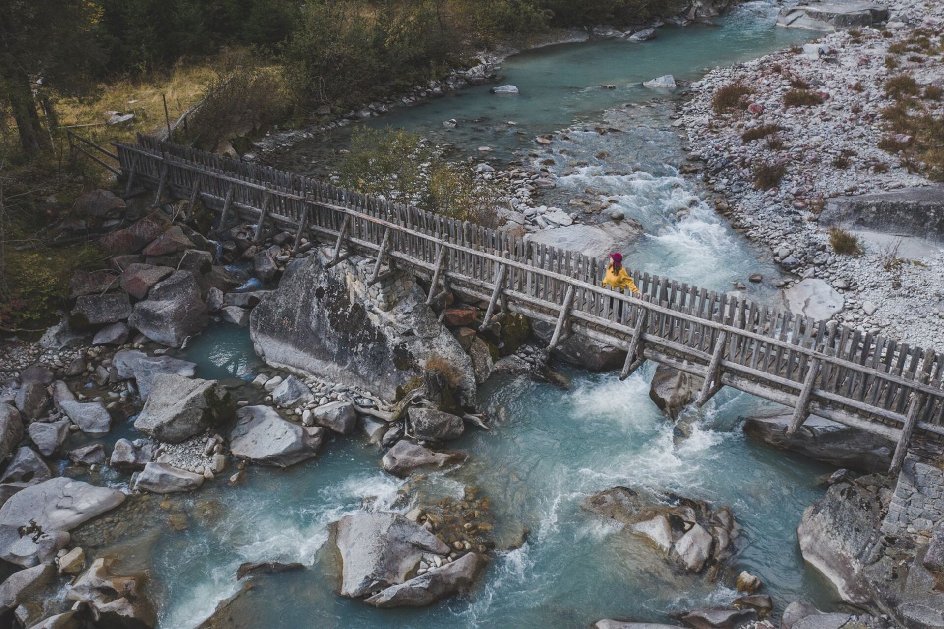 Hochwinkel-Ansicht einer Person in gelber Jacke, die eine charakteristische rustikale Holzbrücke überquert. Darunter fließt der Fluss Sarca mit seinem türkisfarbenen Wasser zwischen Granitblöcken und Felsen. | © Federico Modica, 2019