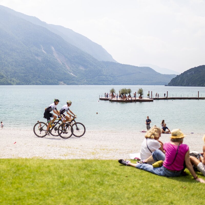 Cyclists riding bikes and people sitting on the beach at Lake Molveno | © Filippo Frizzera, 2019