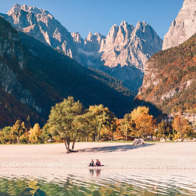 Two people sitting on the beach of Lake Molveno with the Brenta Dolomites behind them in an autumn colours | © Filippo Frizzera, 2019