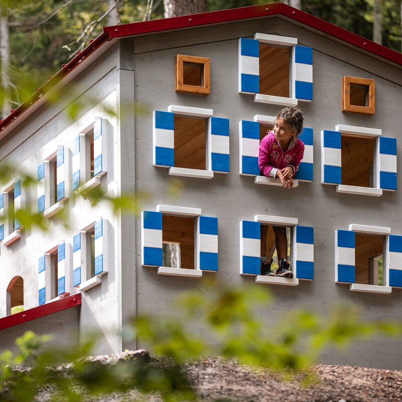 Una bambina si affaccia sorridente dalla finestra di una grande casetta per bambini in legno nel bosco. La struttura ha persiane colorate bianco e blu e un tetto rosso, situata nel parco didattico Sarnacli ad Andalo. | © Oliver Astrologo, 2019