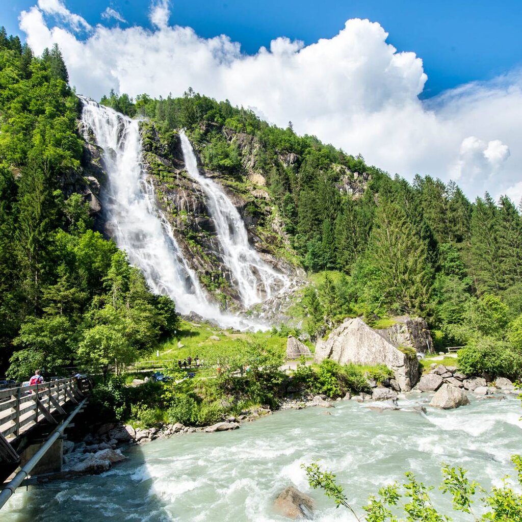 Una vista panoramica delle Cascate Nardis che scendono lungo una parete rocciosa circondata da una fitta foresta verde. In primo piano un torrente impetuoso e un ponte di legno con escursionisti. | © Paolo Bisti, 2015