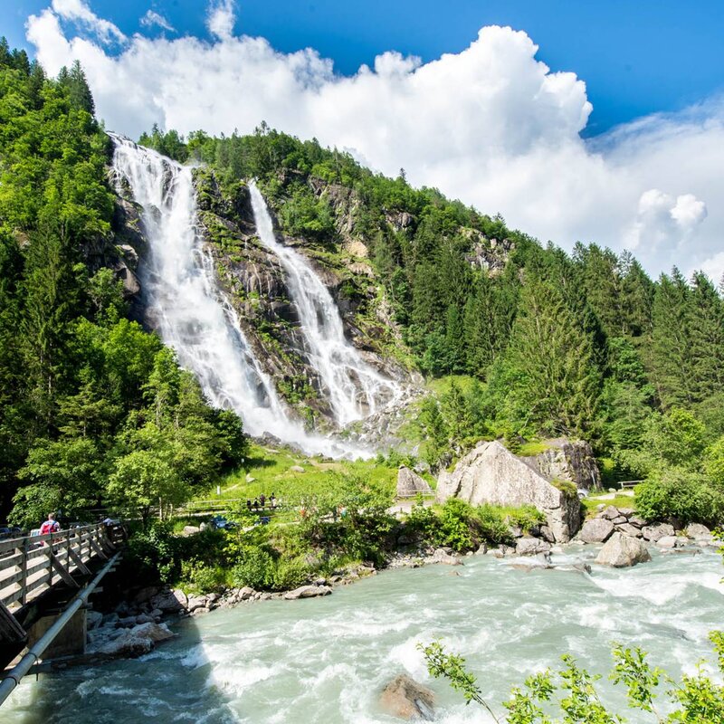 Eine Panorama-Ansicht der Nardis-Wasserfälle, die über eine Felswand inmitten eines dichten grünen Waldes stürzen. Im Vordergrund ein reißender Bach und eine Holzbrücke mit Wanderern. | © Paolo Bisti, 2015