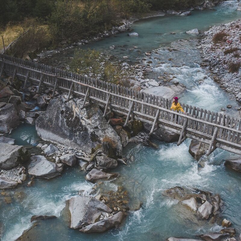 Vista dall'alto di una persona con giacca gialla che attraversa un caratteristico ponte in legno rustico. Sotto scorre il fiume Sarca con le sue acque turchesi tra massi e rocce granitiche. | © Federico Modica, 2019