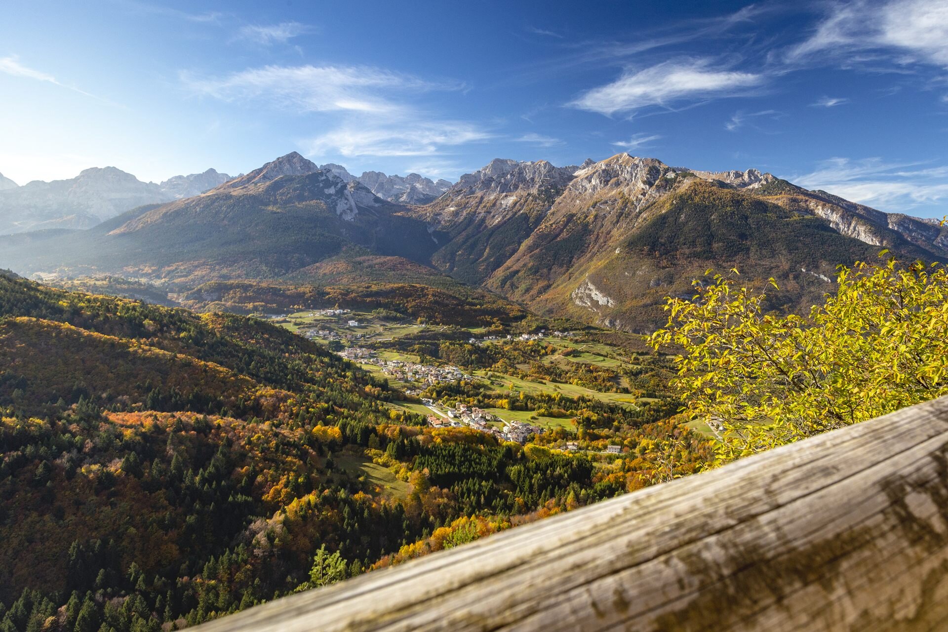 Am Fuße der Brenta-Dolomiten erstreckt sich ein herbstliches Tal mit dem Dorf Cavedago, das sich mit seinen dunklen Häusern an den Talboden schmiegt. Im Vordergrund rahmt ein helles Holzgeländer die Aussicht ein, während ein herbstlich gefärbter Baum auf der rechten Seite für einen Hauch von tiefem Gelb sorgt. Der Himmel ist blau, mit vereinzelten weißen Wolken. | © Filippo Frizzera, 2018