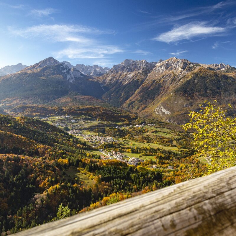 Una valle autunnale si estende ai piedi delle Dolomiti di Brenta, con un villaggio di case dai tetti scuri, Cavedago, adagiato sul fondovalle. In primo piano, una ringhiera di legno chiaro incornicia la vista, con un albero dai colori autunnali che aggiunge un tocco di giallo intenso sulla destra. Il cielo è azzurro, con nuvole bianche sparse. | © Filippo Frizzera, 2018