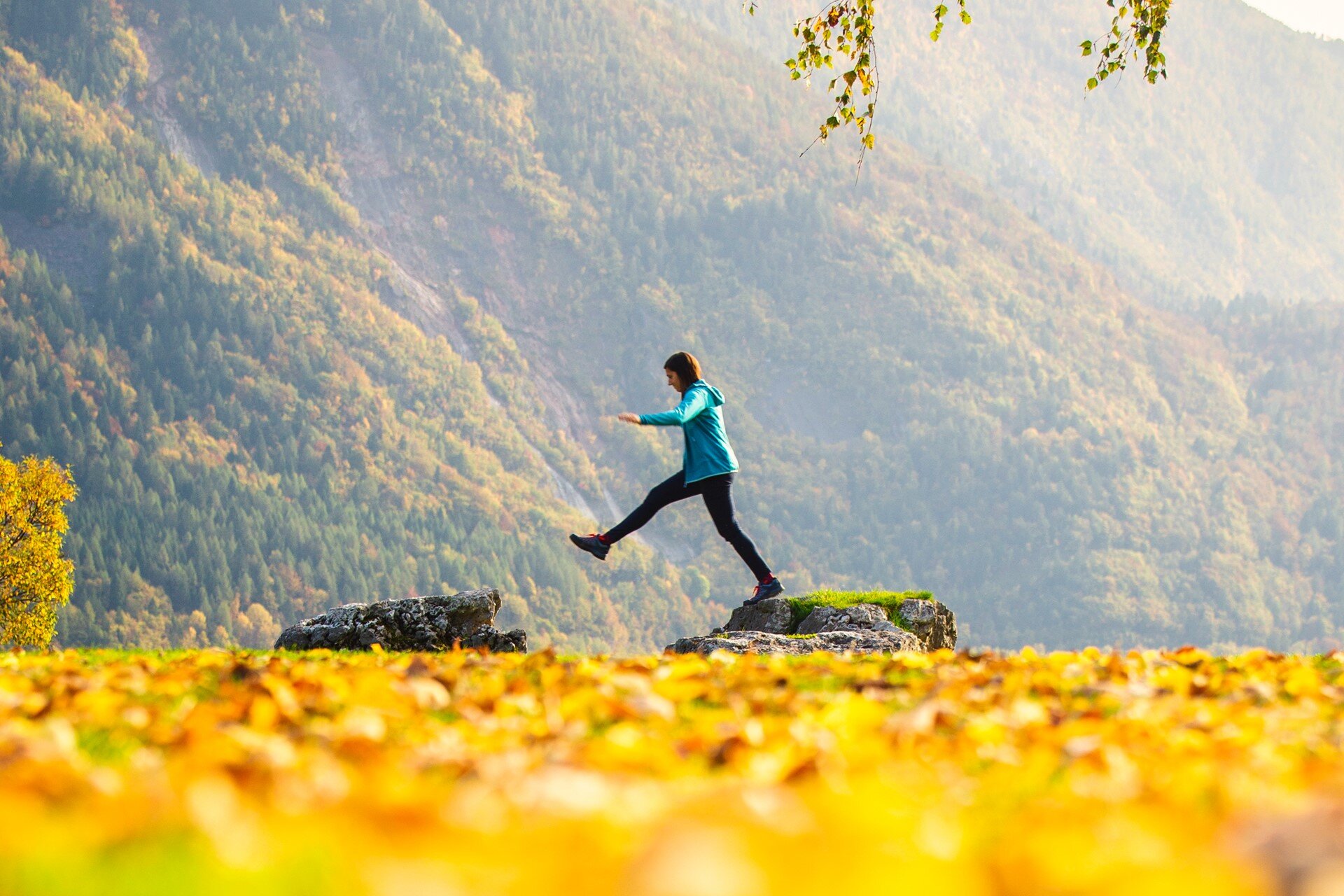 A person is captured mid-air as they leap between rocks on the shore of Lake Molveno in autumn. In the foreground, a carpet of yellow and orange leaves creates a colorful, blurry frame, while a vast mountainside with typical foliage colors stretches out in the background. | © Filippo Frizzera, 2020