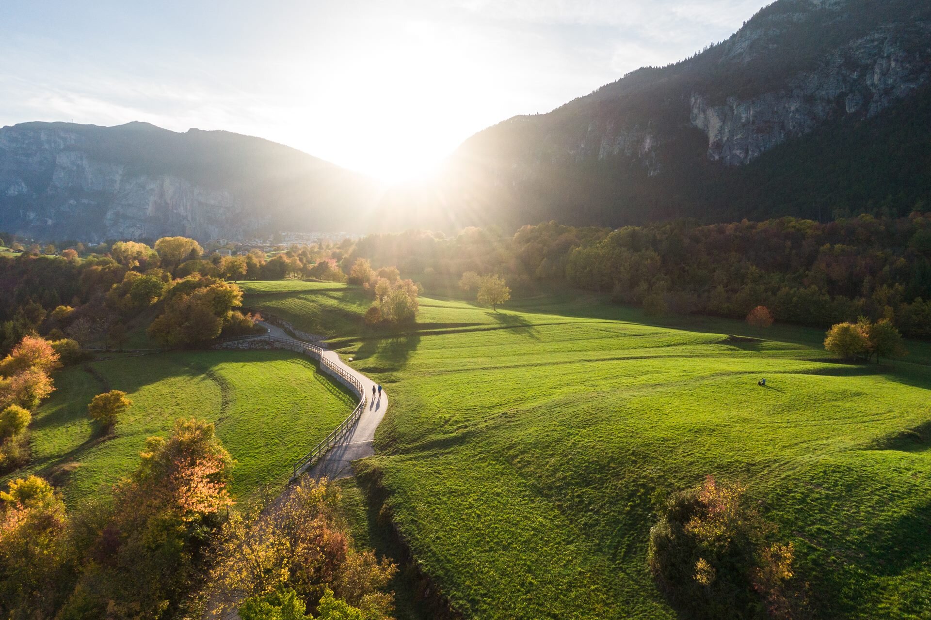 An aerial view of a path winding through a vast green meadow in Fai della Paganella during autumn. The sun is setting behind the mountains, flooding the scene with golden light and casting long shadows, while some trees display the warm colors of the foliage. | © Filippo Frizzera, 2022