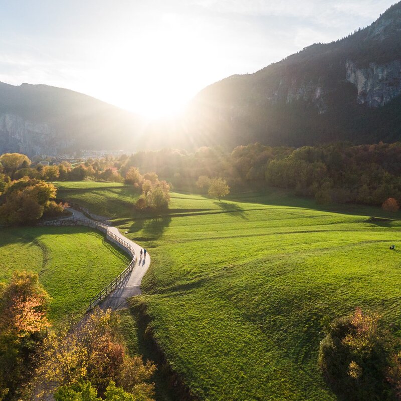Eine Luftaufnahme eines Weges, der sich im Herbst durch eine weite grüne Wiese in Fai della Paganella schlängelt. Die Sonne geht hinter den Bergen unter, überflutet die Szene mit goldenem Licht und wirft lange Schatten, während einige Bäume die warmen Farben des Herbstlaubs zeigen. | © Filippo Frizzera, 2022