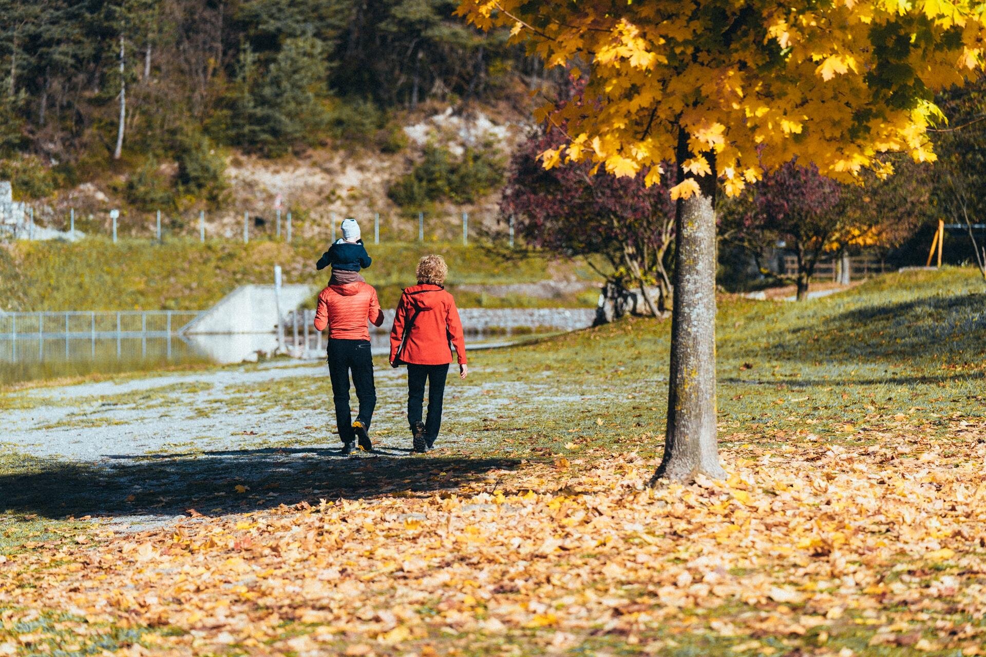 Two adults and a young child are walking along the shores of Lake Nembia on an autumn day. The ground is covered with a carpet of yellow leaves, and one of the adults is carrying the child on their shoulders, in a scene that evokes a peaceful family outing. | © Mirko Perli, 2022