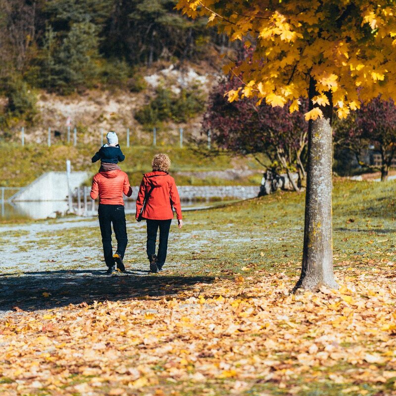 Due adulti e un bambino passeggiano lungo le rive del Lago di Nembia in una giornata d'autunno. Il terreno è coperto da un tappeto di foglie gialle e uno degli adulti porta in spallail bimbo, in una scena che evoca una tranquilla escursione in famiglia. | © Mirko Perli, 2022