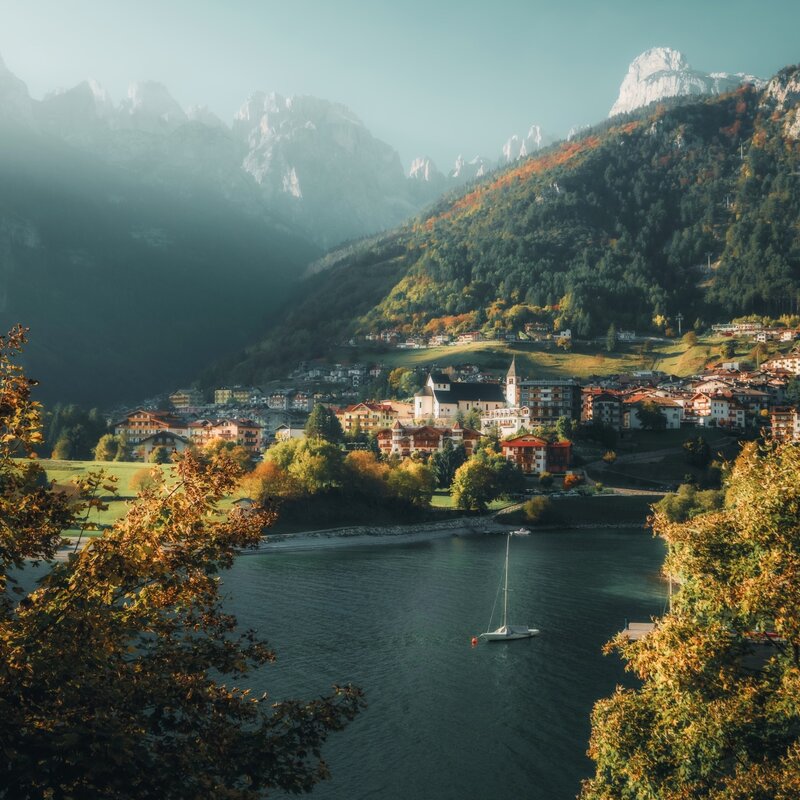 Veduta autunnale del borgo di Molveno affacciato sul lago, incorniciato da alberi dorati e dalle maestose cime delle Dolomiti di Brenta. | © Giulio Gröbert, 2025