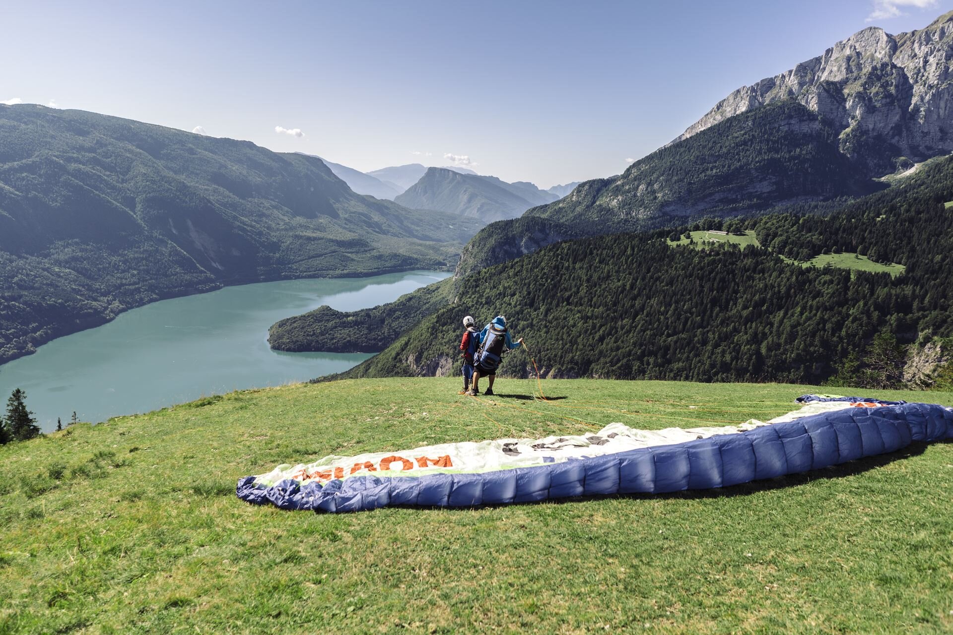 Un istruttore di parapendio sta per alzare la vela del parapendio e decollare dall'altopiano di Pradel per un volo tandem con un'altra persona. Sullo sfondo il lago di Molveno. | © Oliver Astrologo, 2018