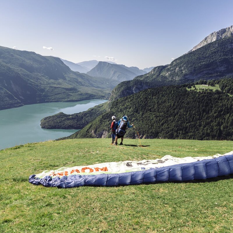 Un istruttore di parapendio sta per alzare la vela del parapendio e decollare dall'altopiano di Pradel per un volo tandem con un'altra persona. Sullo sfondo il lago di Molveno. | © Oliver Astrologo, 2018