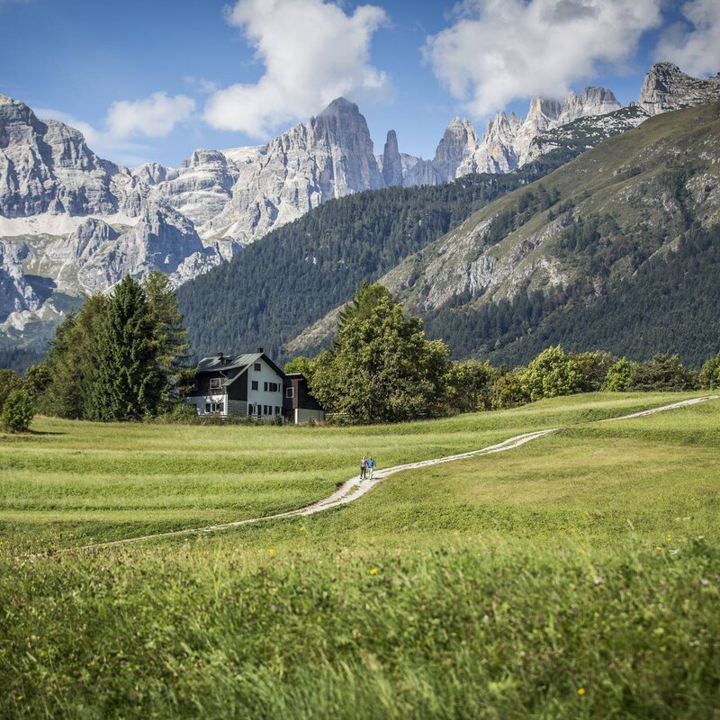 A couple in the distance stroll along the Andalo farmsteads. In the background are the Brenta Dolomites, where the famous Campanil Basso peak stands out. | © Federico Modica, 2016