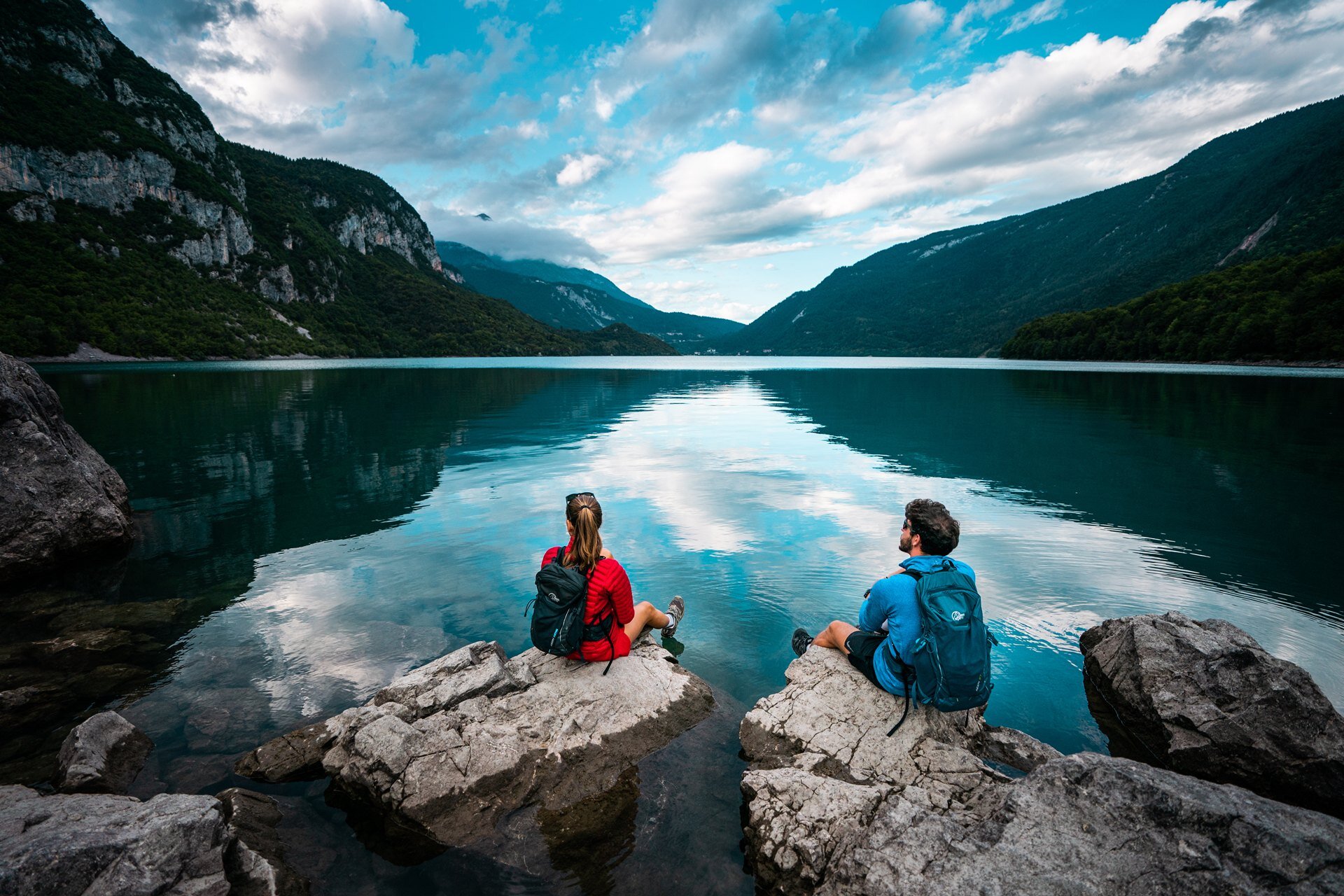 Zwei Wanderer entspannen sich auf den Felsen am Molveno-See und bewundern das perfekte Spiegelbild der Berge auf dem kristallklaren Wasser. Der blaue Himmel und die Wolken schaffen eine heitere und stimmungsvolle Atmosphäre. | © Camilla Pizzini, 2022