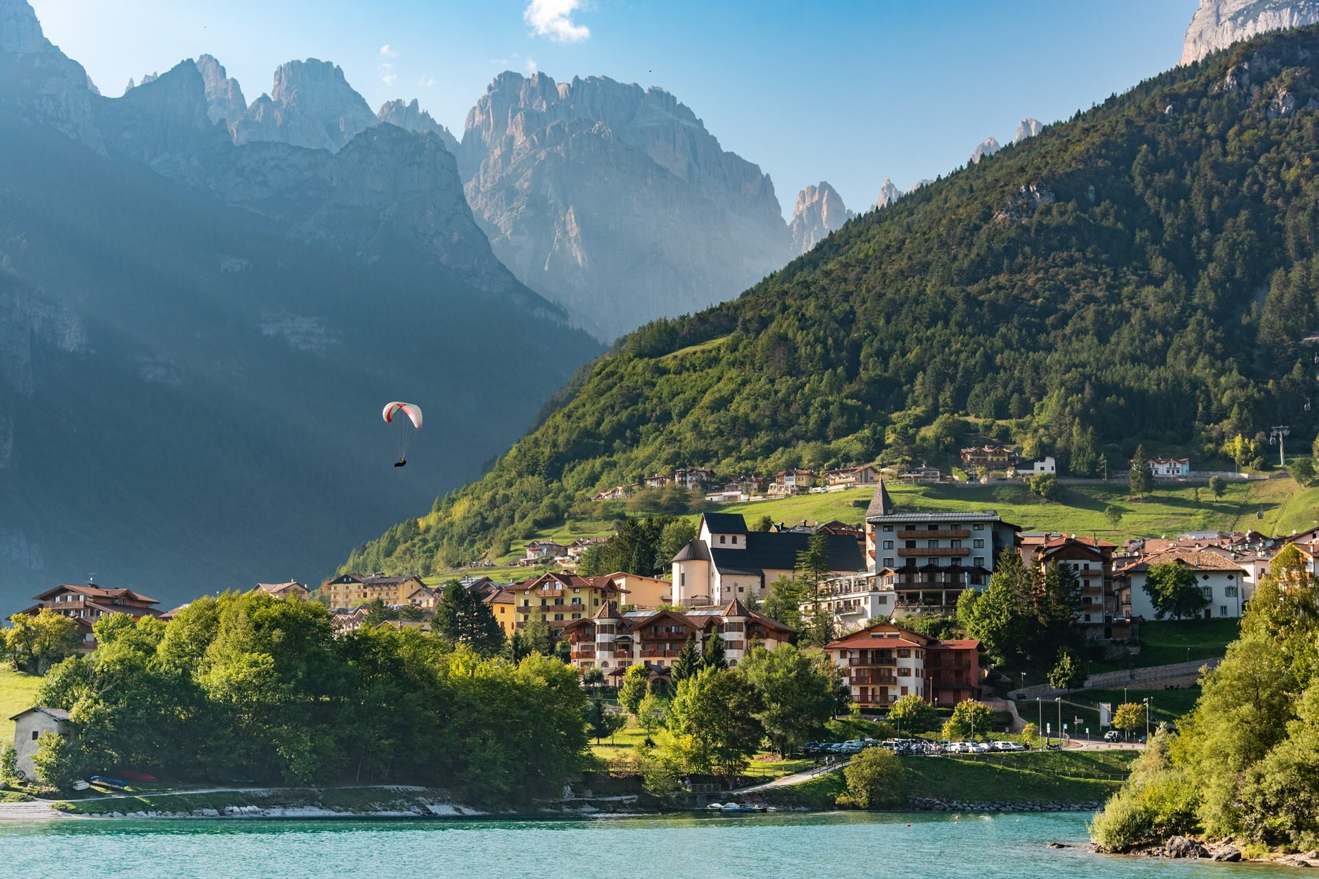 A red and white paraglider flies in the blue sky above the village of Molveno, which lies at the foot of the imposing Brenta Dolomites. | © Veronica Spanu, 2019