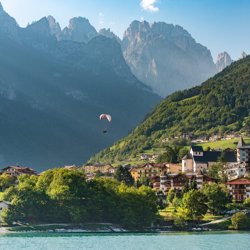Un parapendio rosso e bianco vola nel cielo azzurro sopra il paese di Molveno, che si estende ai piedi delle imponenti Dolomiti di Brenta. | © Veronica Spanu, 2019