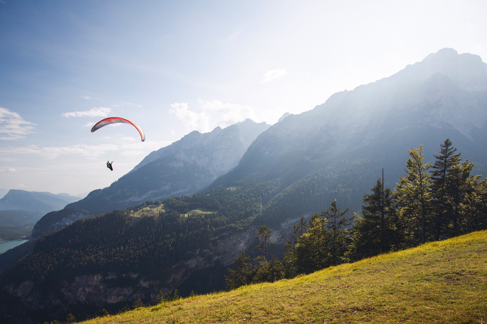 Un parapendio rosso e grigio scende tra le montagne grigie e verdi, stagliandosi contro un cielo azzurro chiaro. L'orizzonte è alto, con la cima di una montagna che si erge sulla destra e un piccolo scorcio del lago di Molveno in lontananza a sinistra. In basso a destra, un pendio erboso chiaro completa il paesaggio. | © Filippo Frizzera, 2019