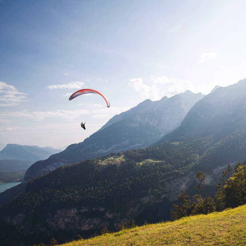 Un parapendio rosso e grigio scende tra le montagne grigie e verdi, stagliandosi contro un cielo azzurro chiaro. L'orizzonte è alto, con la cima di una montagna che si erge sulla destra e un piccolo scorcio del lago di Molveno in lontananza a sinistra. In basso a destra, un pendio erboso chiaro completa il paesaggio. | © Filippo Frizzera, 2019