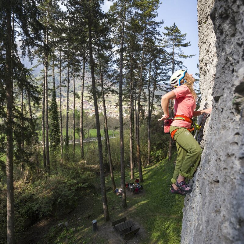 Una ragazza arrampica in falesia in un bosco. Indossa casco e scarpe da climber, panataloni verdi e una t-shirt rosa. Tra gli alberi si intravede, sullo sfondo, il borgo di San Lorenzo Dorsino. | © Filippo Frizzera, 2017