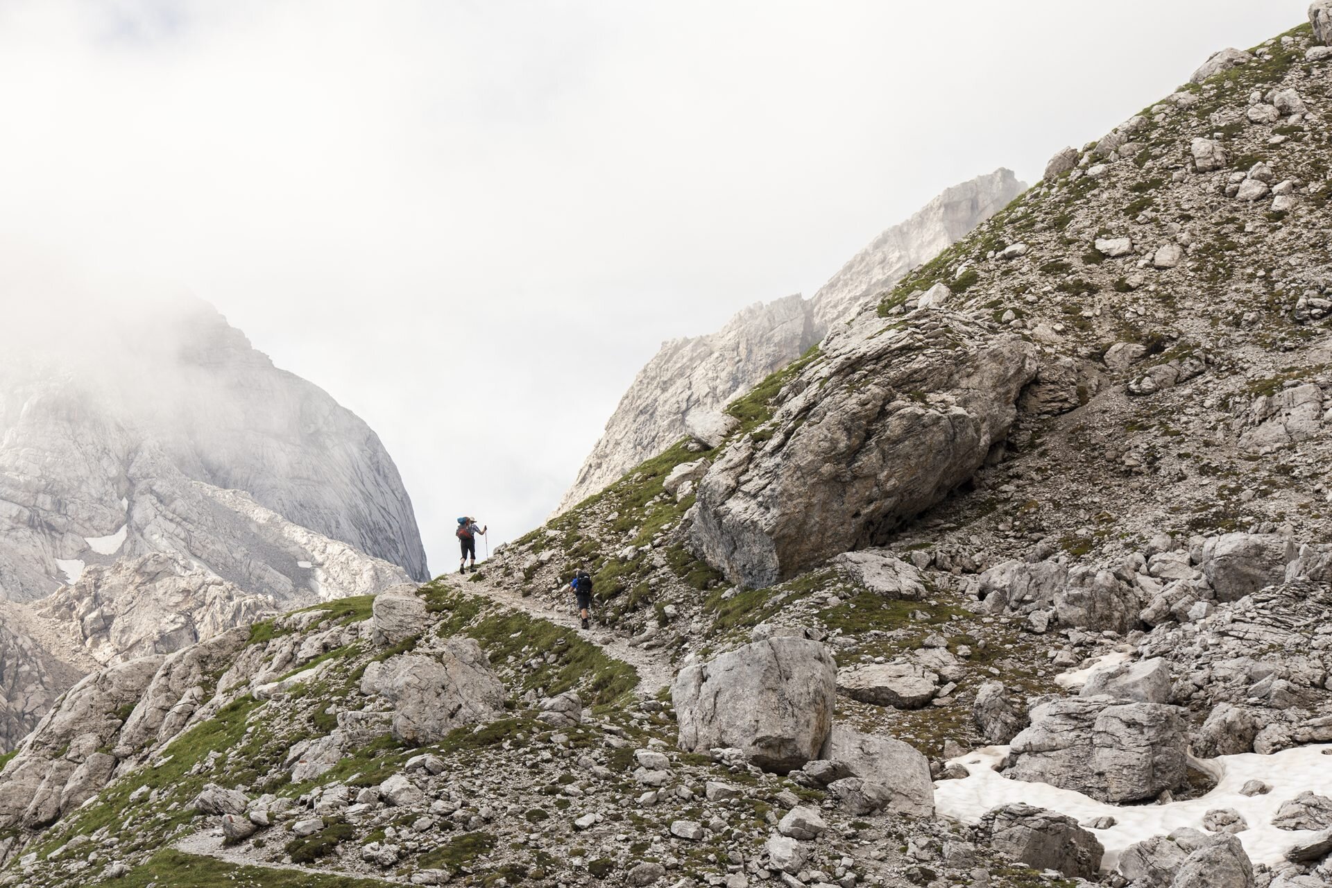 Zwei Wanderer, von hinten und in der Ferne gesehen, gehen auf einem schmalen, gewundenen Pfad einen steilen Felshang der Brenta-Dolomiten hinauf. Die Landschaft ist geprägt von großen grauen Felsbrocken und spärlicher Vegetation. Im Hintergrund erheben sich imposante, teilweise nebelverhangene Berge unter einem wolkenverhangenen Himmel. | © Filippo Frizzera, 2019