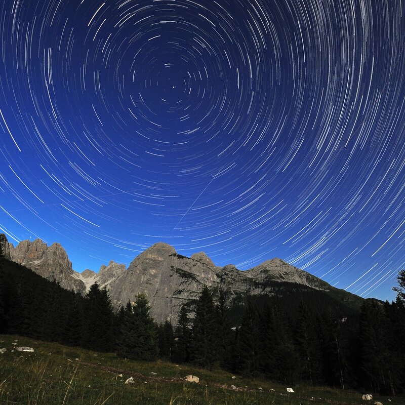 Cielo notturno stellato sopra un paesaggio montano. Le stelle appaiono come scie luminose circolari attorno a un punto centrale, effetto dovuto a una lunga esposizione fotografica che cattura il movimento apparente della volta celeste. In basso, si stagliano le sagome scure delle montagne e degli alberi contro il cielo blu intenso. | © Claudio Donini, 2011
