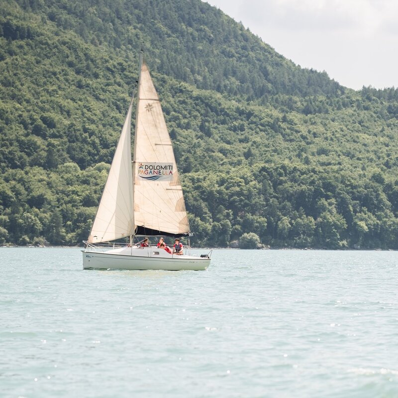 Una barca a vela bianca naviga su un lago calmo, con alcune persone a bordo. Sullo sfondo, un'ampia collina ricoperta da una fitta foresta verde scende fino alla riva del lago. | © Filippo Frizzera, 2024