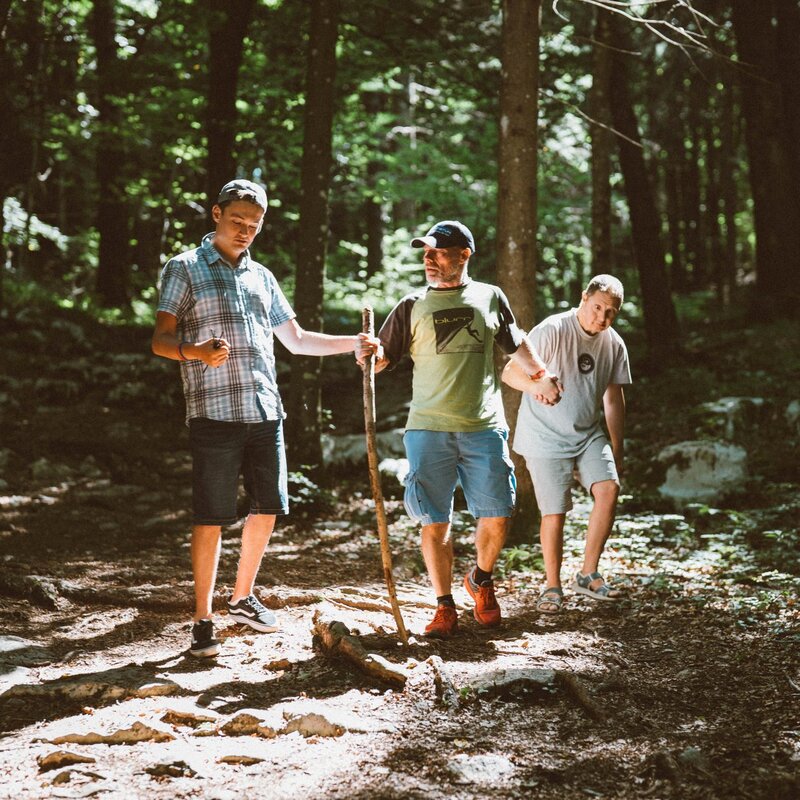 Tre uomini camminano lungo un sentiero sterrato nel bosco del Parco del Respiro a Fai della Paganella. L'uomo al centro utilizza un lungo ramo come bastone da appoggio; alla sua destra, un compagno lo tiene per mano per accompagnarlo nel cammino, mentre alla sua sinistra un altro giovane procede al suo fianco. La luce del sole filtra tra le chiome degli alberi creando zone di luce e ombra sul terreno. | © Mirko Perli, 2025