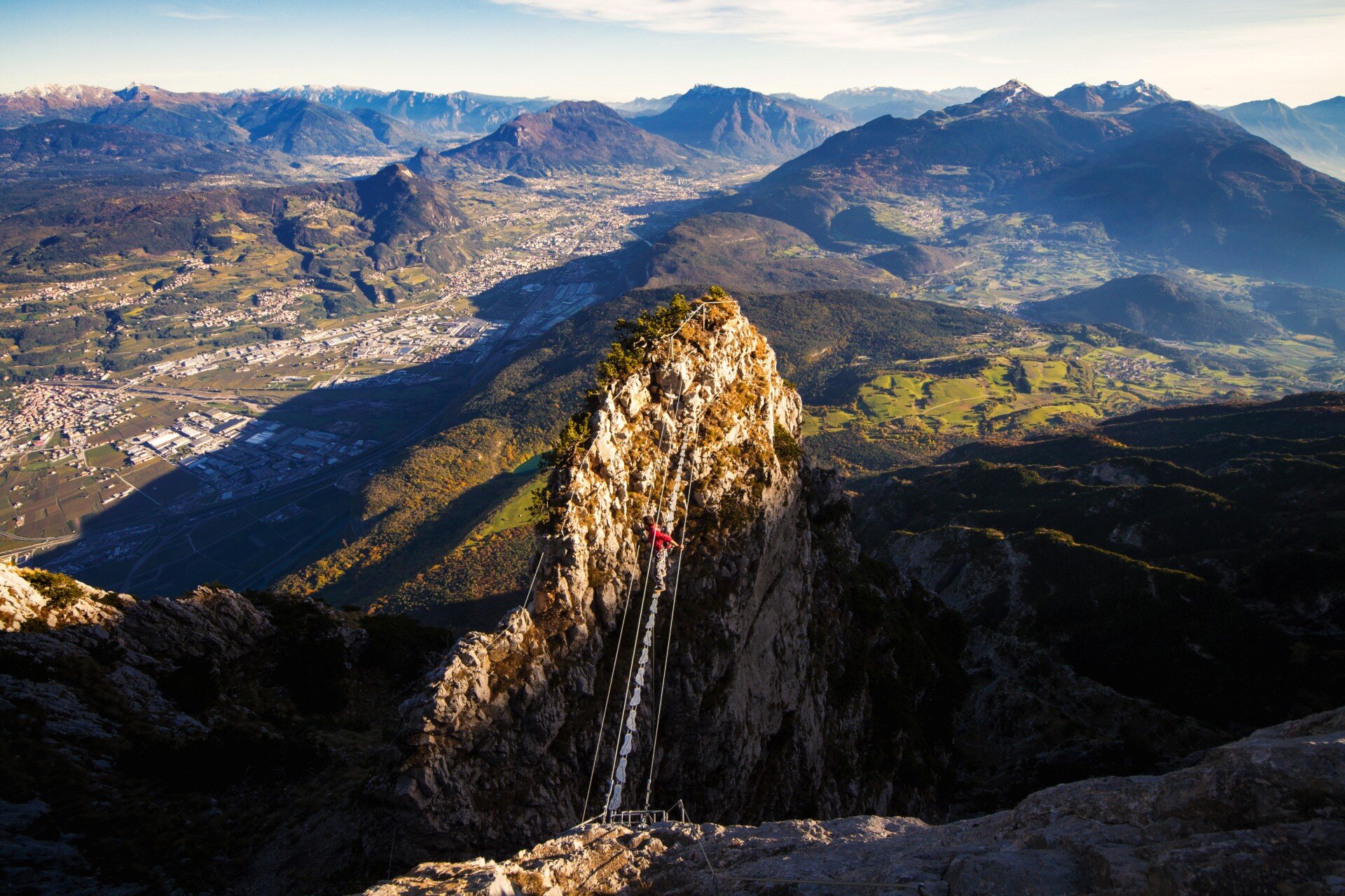A person crosses a suspension bridge made of steel cables strung between two rocky pinnacles. In the background, a vast aerial view of a valley with towns, fields, and distant mountain ranges. | © Filippo Frizzera, 2015