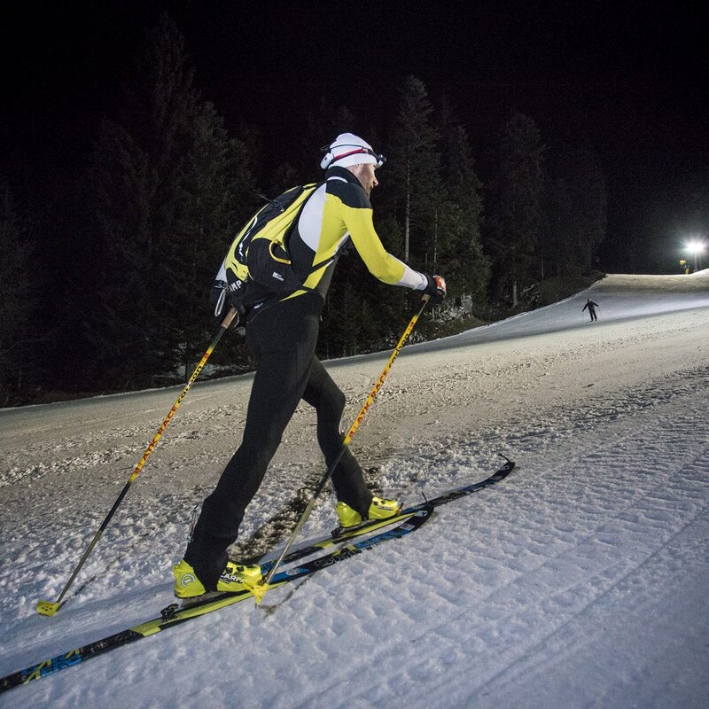  L'immagine mostra uno sciatore di sci alpinismo che risale una pista innevata di notte. Indossa un abbigliamento sportivo giallo e nero, un cappello bianco e una lampada frontale. Lo sciatore è equipaggiato con zaino, bastoncini e sci con pelli di foca per la salita. Sullo sfondo, la pista è illuminata da alcune luci artificiali, mentre l'area circostante è avvolta nell'oscurità, con alberi che si stagliano contro il cielo notturno. Un altro sciatore è visibile in lontananza | © Filippo Frizzera, 2018