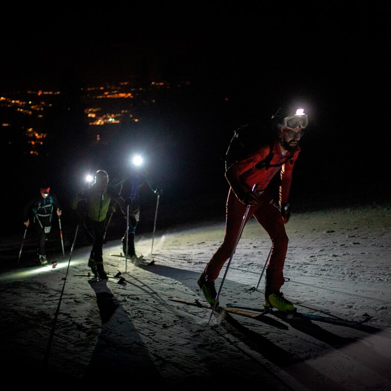 L'immagine mostra un gruppo di sciatori che affronta una salita su neve in notturna, illuminati solo dalle luci dei loro caschi e dall'illuminazione artificiale sullo sfondo. La scena è molto scura, con un contrasto netto tra le figure degli sciatori e il paesaggio innevato. Lo sciatore in primo piano indossa una tuta rossa e occhiali protettivi, mentre altri due sciatori seguono leggermente distanti, anch'essi con lampade frontali accese. Sullo sfondo, si vedono le luci della città, che brillano nel buio della notte. | © Filippo Frizzera, 2019