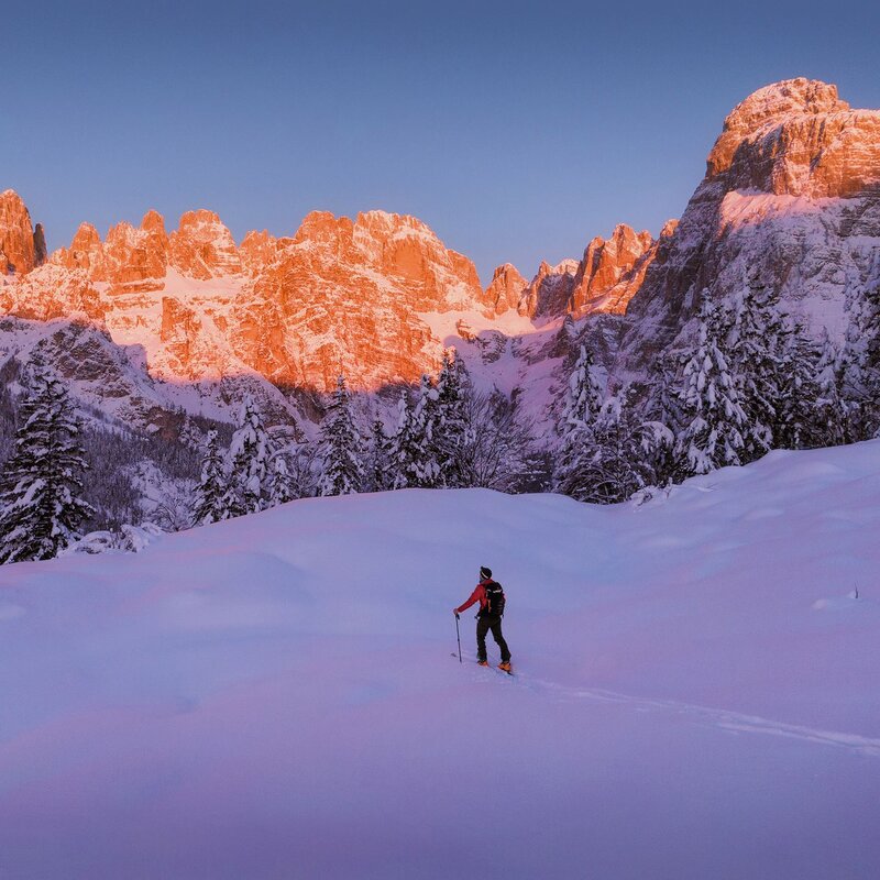 Das Bild zeigt eine Berglandschaft im Morgengrauen, wobei die Gipfel der Brenta-Dolomiten von einem warmen, rötlichen Licht, der Enrosadira, beleuchtet werden. Im Vordergrund fährt ein einsamer Skifahrer über eine Fläche unberührten Schnees, umgeben von schneebedeckten Bäumen. Der Himmel ist klar und von einem intensiven Blau. Die Atmosphäre ist ruhig und still und erinnert an die Schönheit und Weite der winterlichen Alpenwelt. | © Filippo Frizzera, 2019