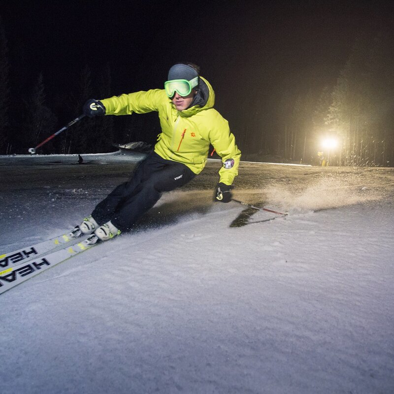 This photo shows a skier skiing down a snowy slope at night, wearing a yellow jacket, dark pants and ski goggles. The lighting comes from a powerful spotlight behind him, illuminating the snow. The skier is in a dynamic, prone position. | © Filippo Frizzera, 2018