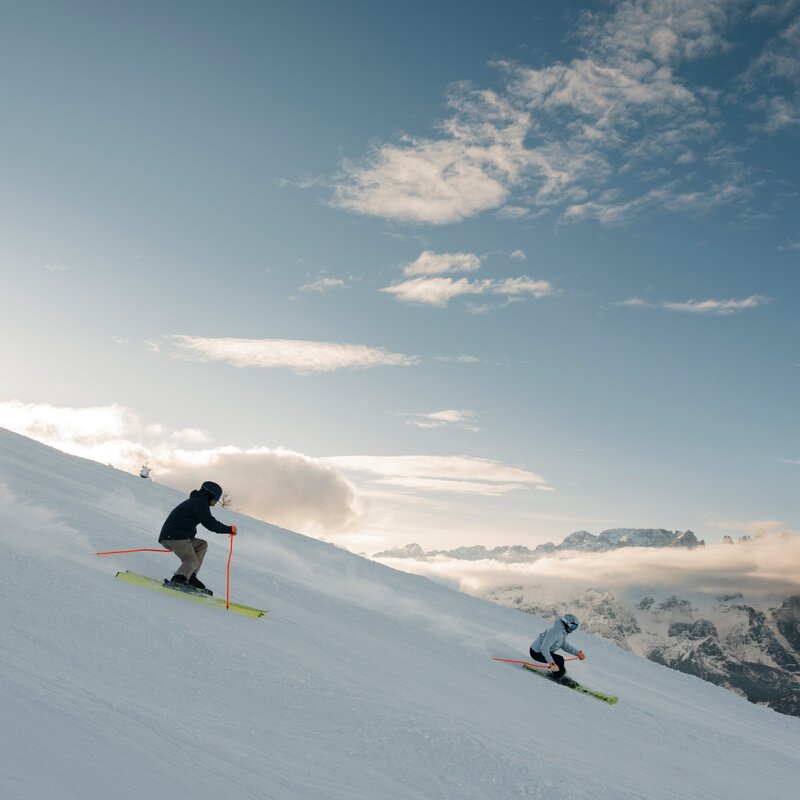 Two skiers carving fast down a wide, sunny slope. The sky is blue with low clouds, and the snow-capped Brenta Dolomites are visible in the distance. | © Federico Modica, 2026