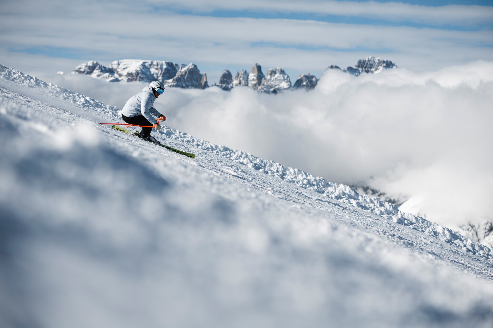 A skier in a light blue jacket carves a turn on the snow. Behind them, the rocky peaks of the Brenta Dolomites rise above a thick layer of white clouds. | © Federico Modica, 2026