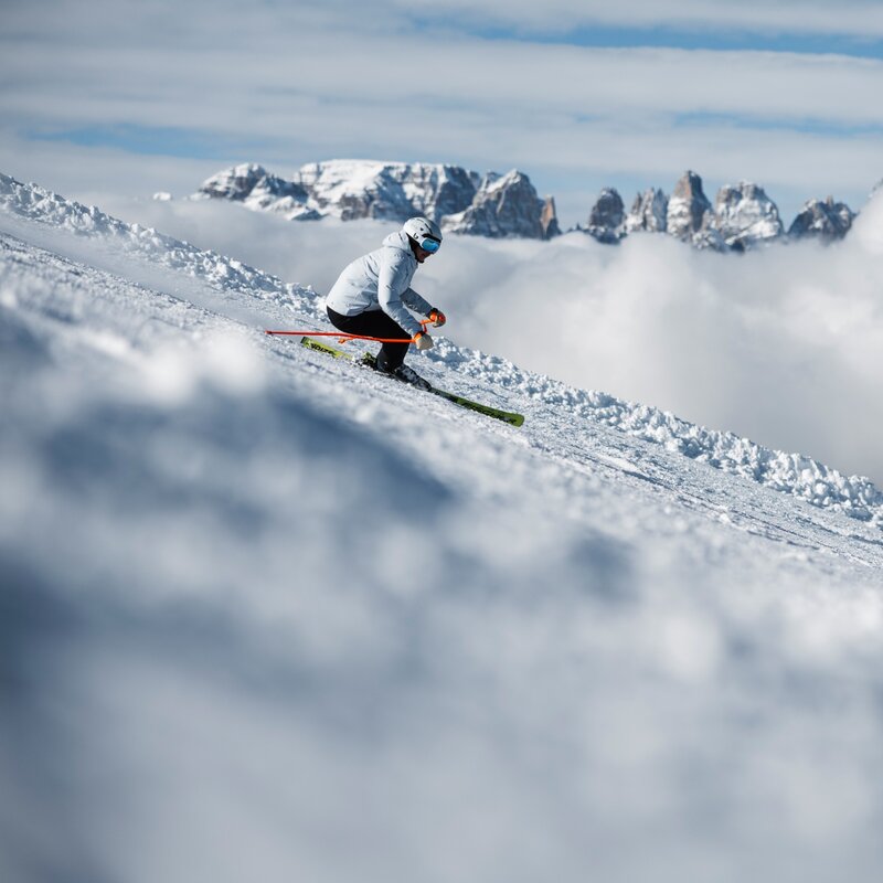 Una sciatrice con giacca azzurra effettua una curva sulla neve. Alle sue spalle, le cime rocciose delle Dolomiti di Brenta si ergono sopra uno strato compatto di nuvole bianche. | © Federico Modica, 2026
