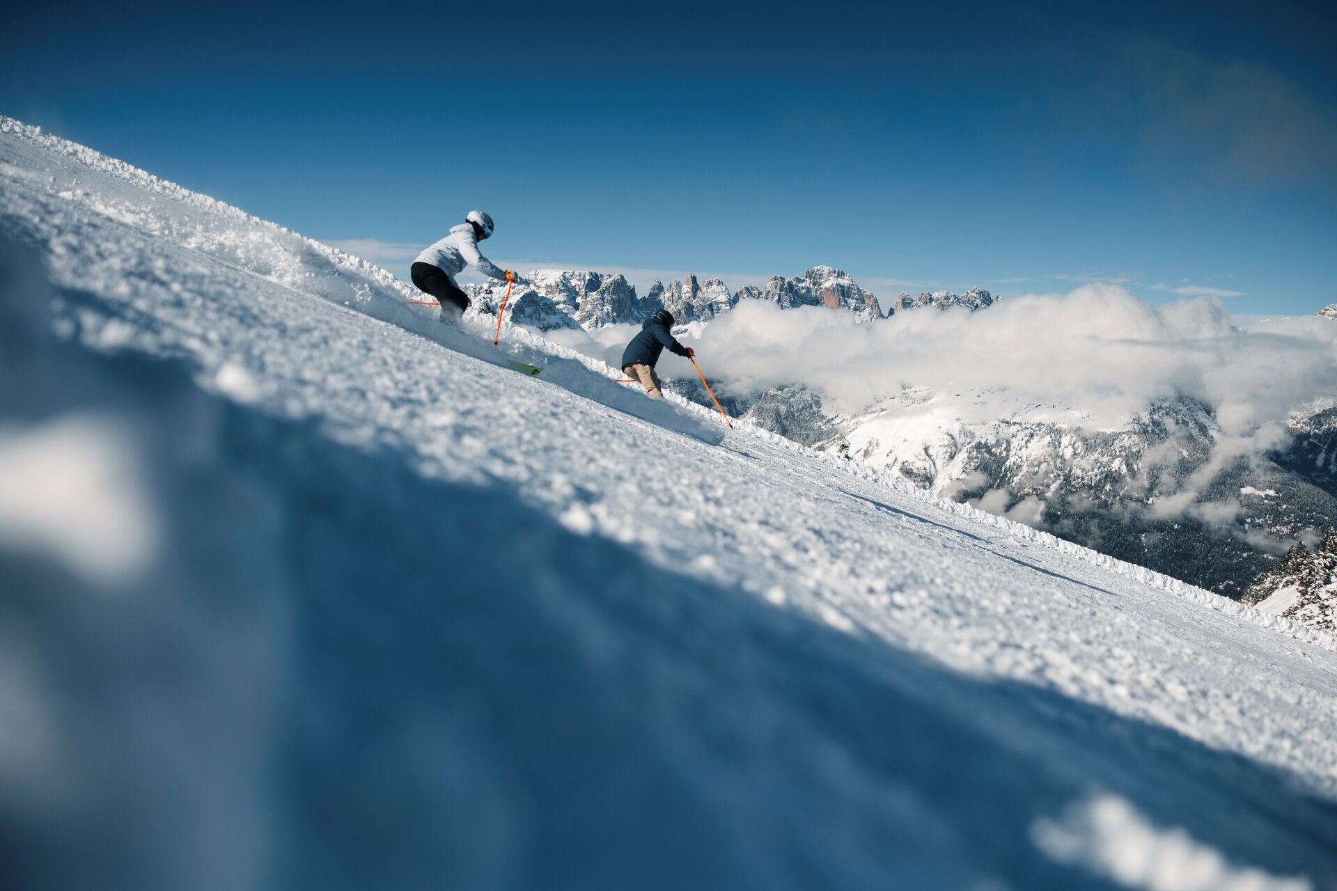 Two skiers descend a steep, snow-covered slope, kicking up fresh snow as they go. In the background, under a clear blue sky, rocky mountain peaks emerge from a dense layer of white clouds. | © Federico Modica, 2026