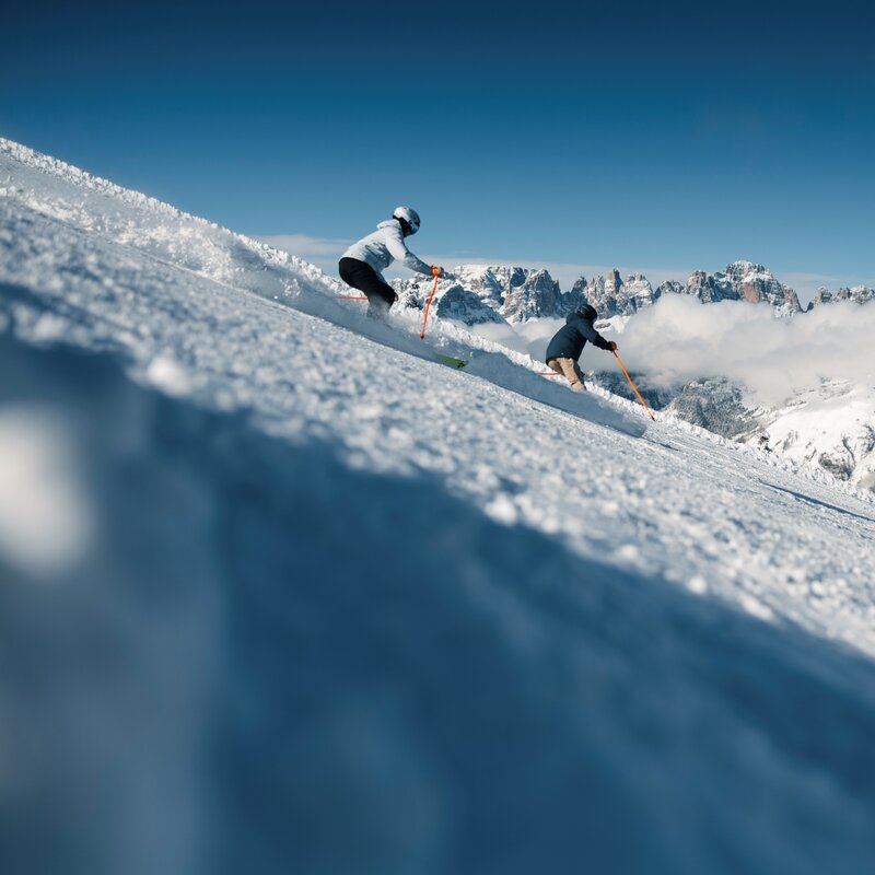 Two skiers descend a steep, snow-covered slope, kicking up fresh snow as they go. In the background, under a clear blue sky, rocky mountain peaks emerge from a dense layer of white clouds. | © Federico Modica, 2026
