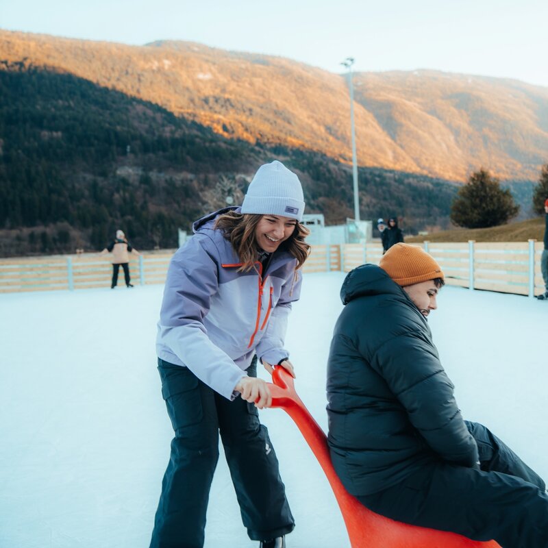 A smiling young woman in a light jacket and white beanie pushes a young man sitting on a red skating aid on an outdoor ice rink. In the background, other people skate near a wooden fence, surrounded by sunlit mountains. | © Jo Sirigu, 2026