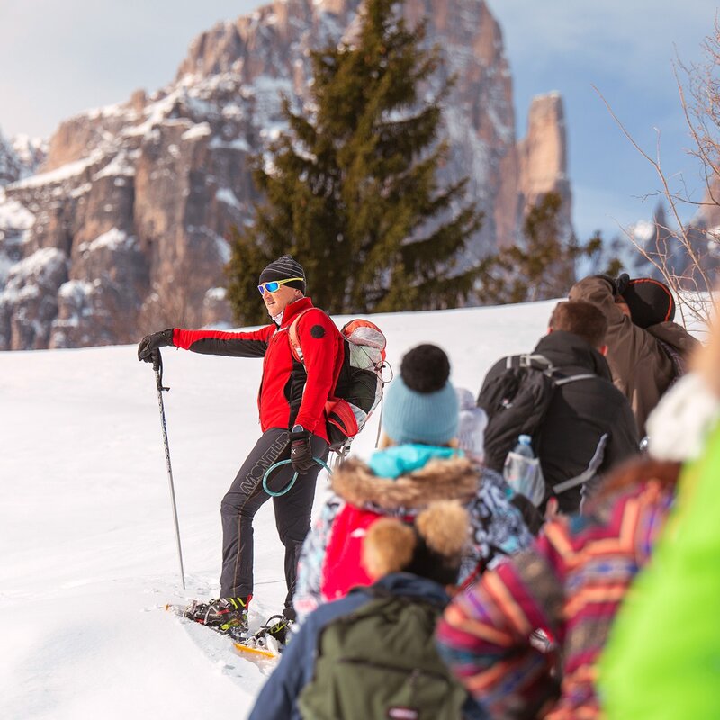 Das Bild zeigt eine Gruppe von Menschen in einer Reihe auf einem verschneiten Weg. Im Vordergrund ist ein rot gekleideter Führer mit Sonnenbrille zu sehen, der die Gruppe mit Schneeschuhstöcken anspricht. Im Hintergrund sind Bäume und die schneebedeckten Brenta-Dolomiten zu sehen. | © Filippo Frizzera, 2019