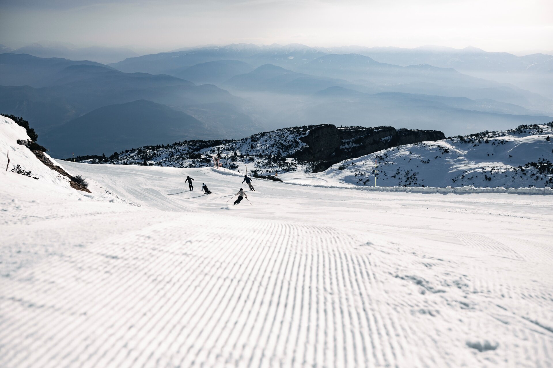 Vista da una pista da sci con neve battuta a righe in primo piano. Quattro sciatori scendono a distanza media. Sullo sfondo, montagne innevate e valli sfumate dalla foschia sotto un cielo chiaro. | © Federico Modica, 2026