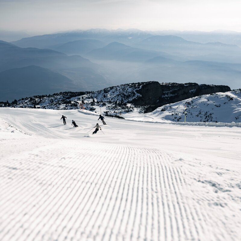 Blick von einer Skipiste mit präpariertem, geriffeltem Schnee im Vordergrund. Vier Skifahrer fahren in mittlerer Entfernung ab. Im Hintergrund verschneite Berge und dunstige Täler unter einem klaren Himmel. | © Federico Modica, 2026