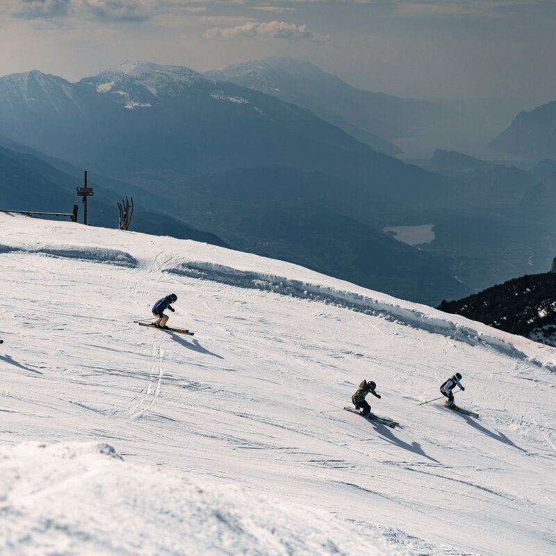 Quattro sciatori in discesa su una pista della Ski Area Paganella. In primo piano neve battuta; sullo sfondo le valli del Trentino e il Lago di Garda visibile tra le montagne. | © Federico Modica, 2026