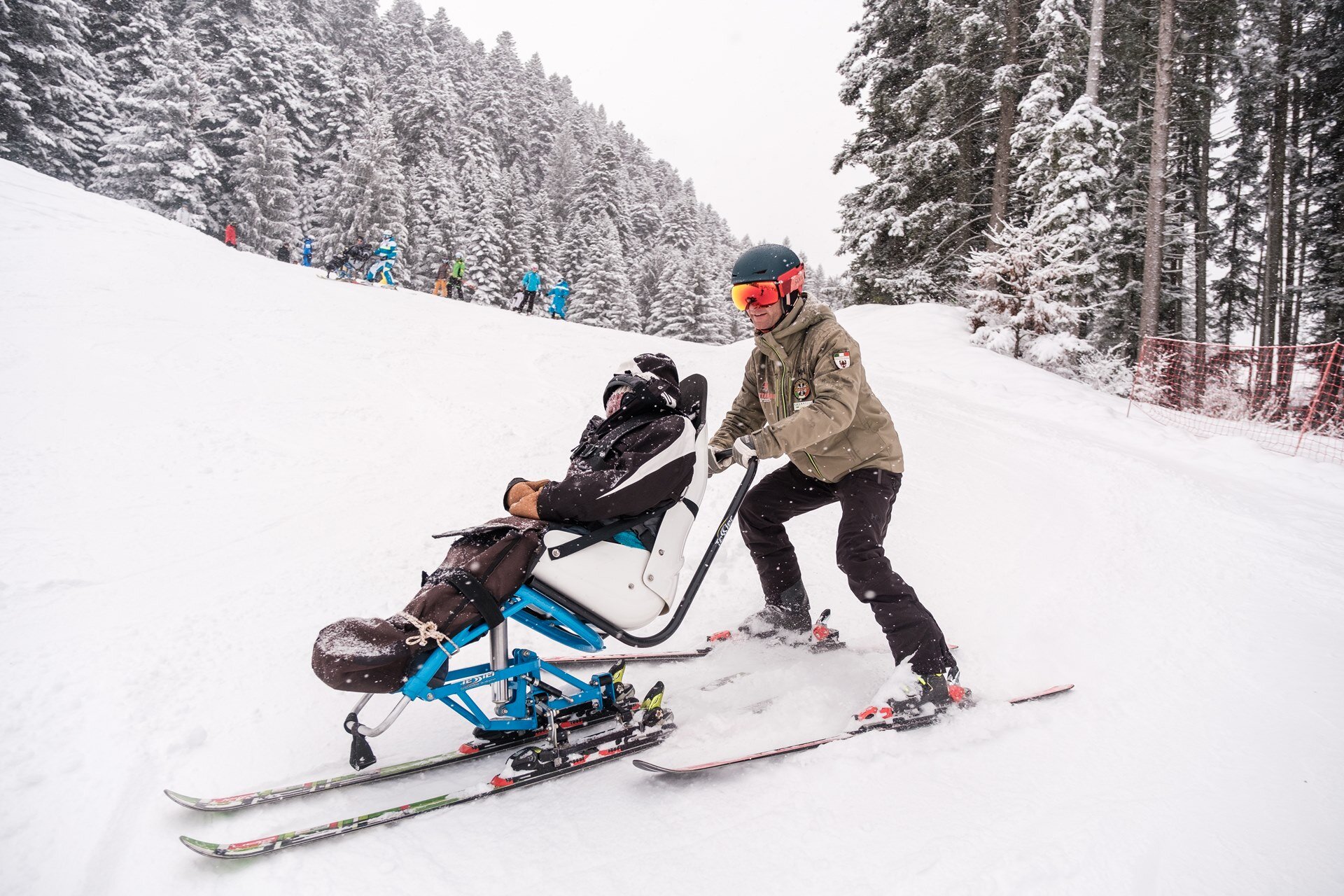 Un maestro di sci scende lungo una pista portando un monosci, un ausilio per persone con disabilità. Nevica molto e la neve che cade copre in parte il maestro di sci e la persona sul monosci. | © Filippo Frizzera, 2023