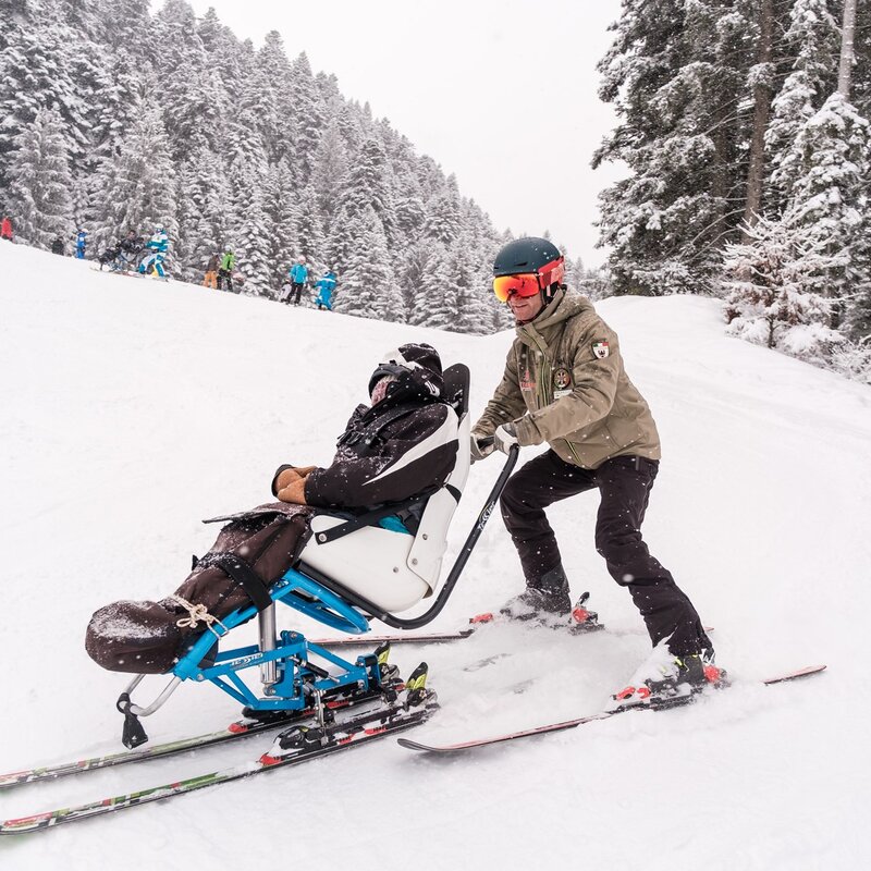 Un maestro di sci scende lungo una pista portando un monosci, un ausilio per persone con disabilità. Nevica molto e la neve che cade copre in parte il maestro di sci e la persona sul monosci. | © Filippo Frizzera, 2023