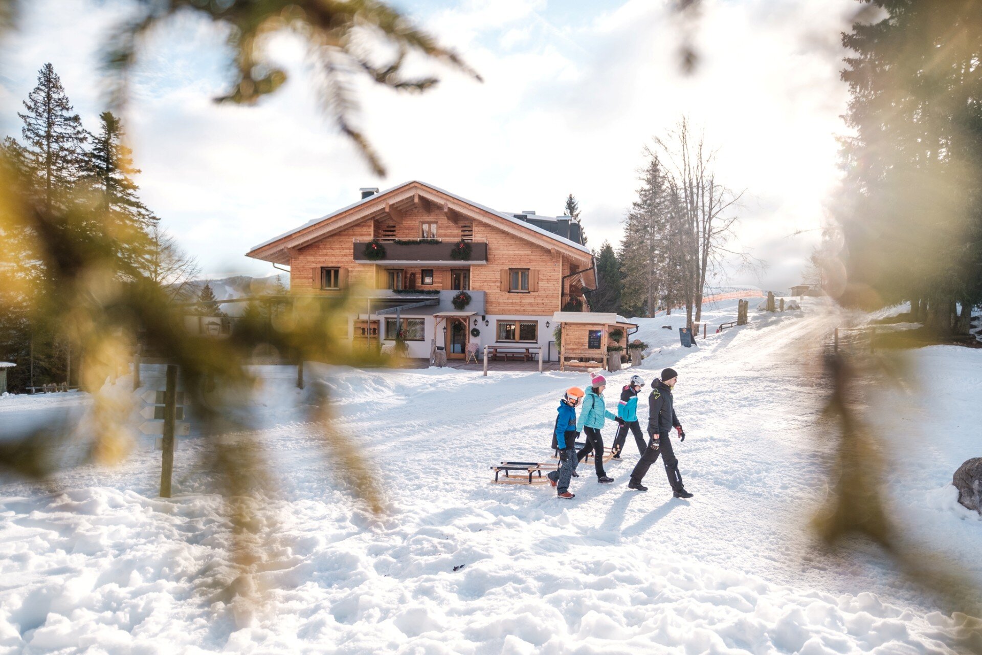 A family of two adults and two children walks on the snow pulling wooden sleds. The scene is framed by blurred fir branches and takes place in front of a large traditional wooden chalet, illuminated by warm, diffused sunlight. | © Filippo Frizzera, 2022