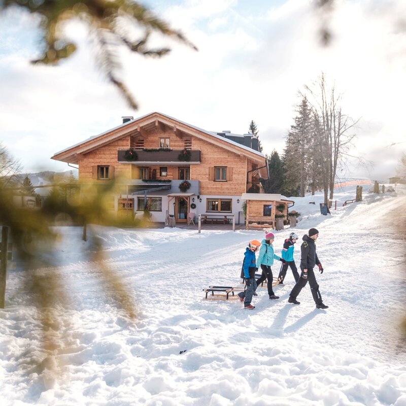 Una famiglia composta da due adulti e due bambini cammina sulla neve tirando slittini di legno. La scena è incorniciata da rami di abete sfocati e si svolge davanti a un grande chalet tradizionale in legno, illuminato da una luce solare calda e diffusa. | © Filippo Frizzera, 2022