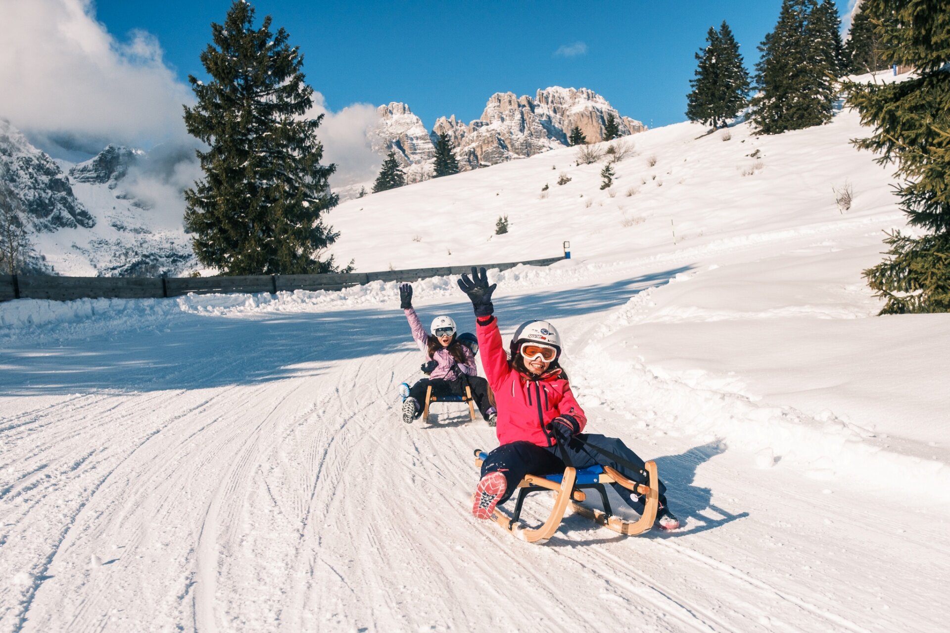 Two children descend on sleds along a groomed snowy track. The child in the foreground smiles and raises a hand to wave. In the background, green fir trees and rocky mountain peaks stand out against a blue sky with clouds. | © Filippo Frizzera, 2026